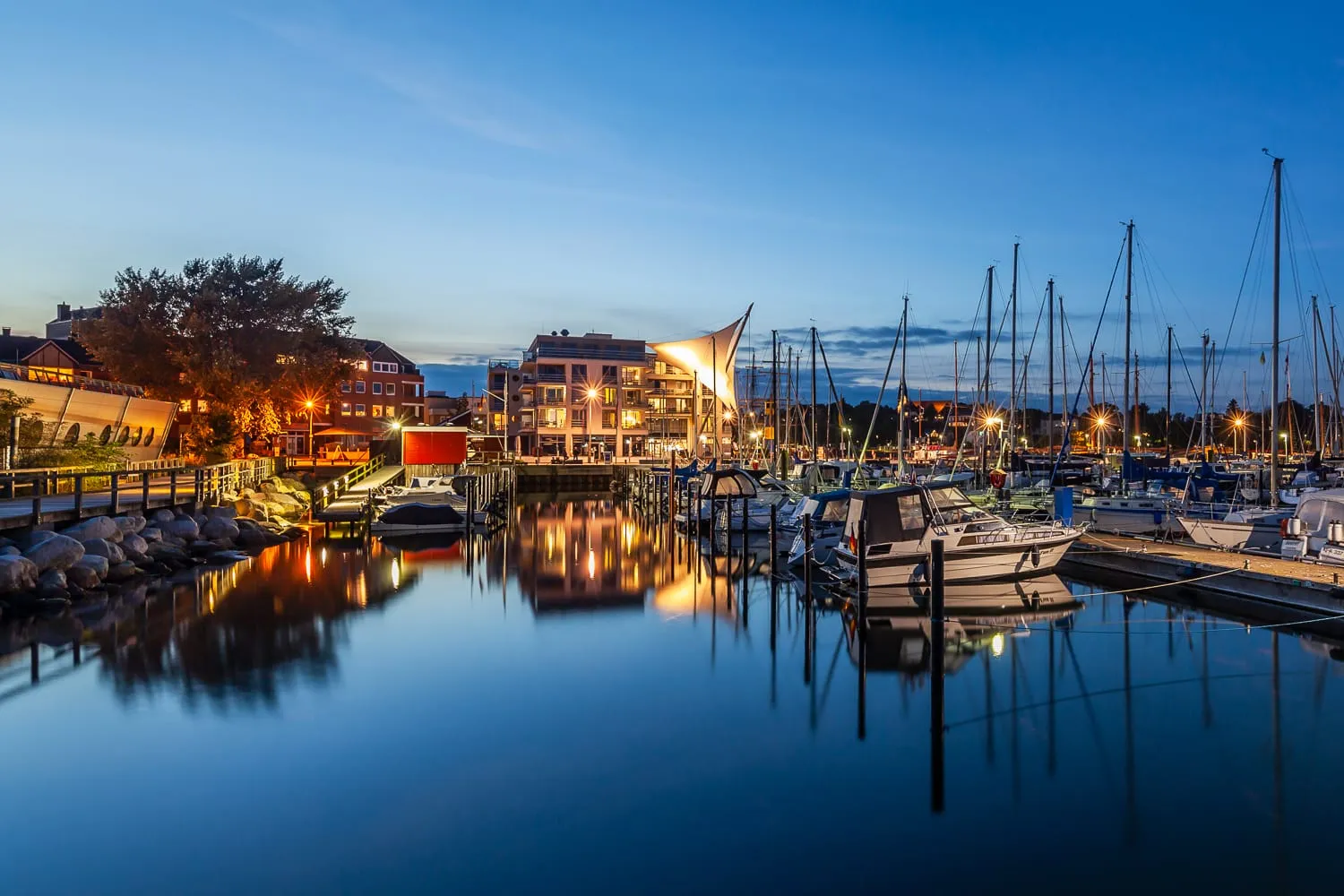 Der Yachthafen in der blauen Stunde, Lichter und Masten spiegeln sich, hinten das Haus mit den Segeln