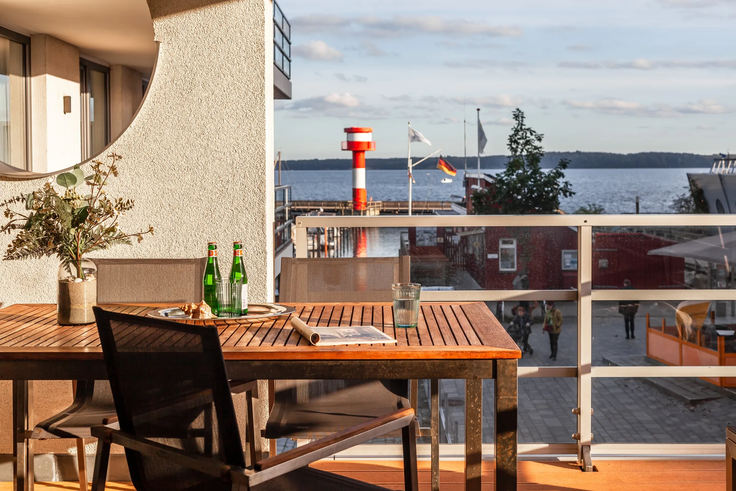 Teaktisch mit Wasserflaschen auf der Loggia, dahinter der rot-weiße Leuchtturm und die Bucht