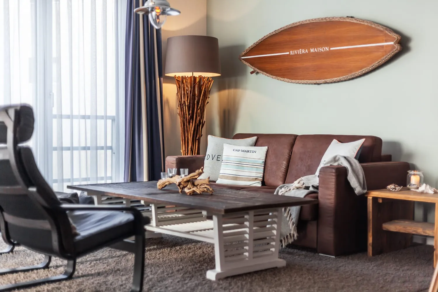 Sofa, Ledersessel und Couchtisch vor den großen Fenstern, an der Wand das Surfbrett