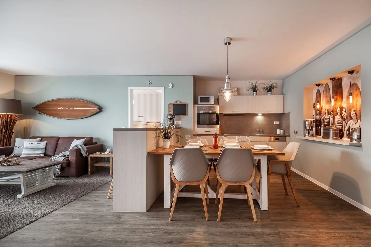 Blick über den Wohnraum: Sitzecke mit Surfbrett an der Wand, Küchentresen und gedeckter Esstisch